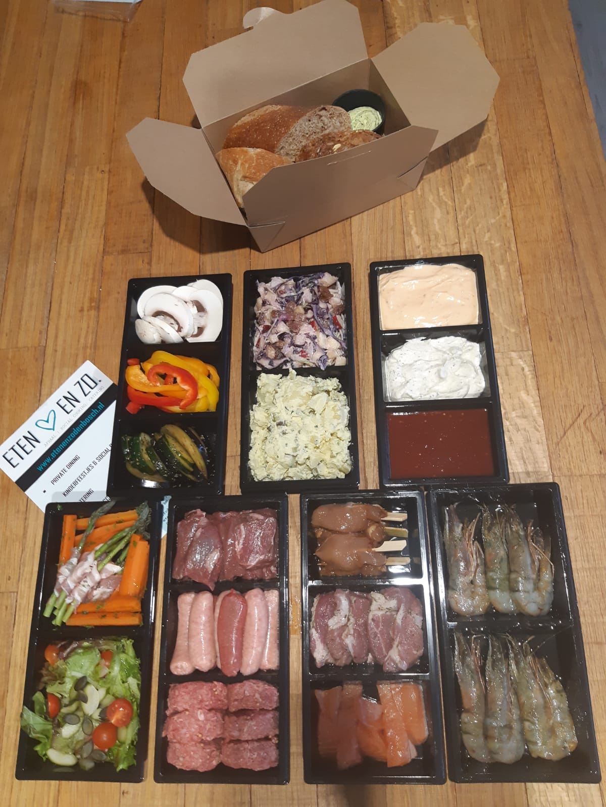 Een assortiment rauw vlees, vis, verse groenten, salades en dipsauzen voor een barbecue thuis, uitgestald op een houten tafel.