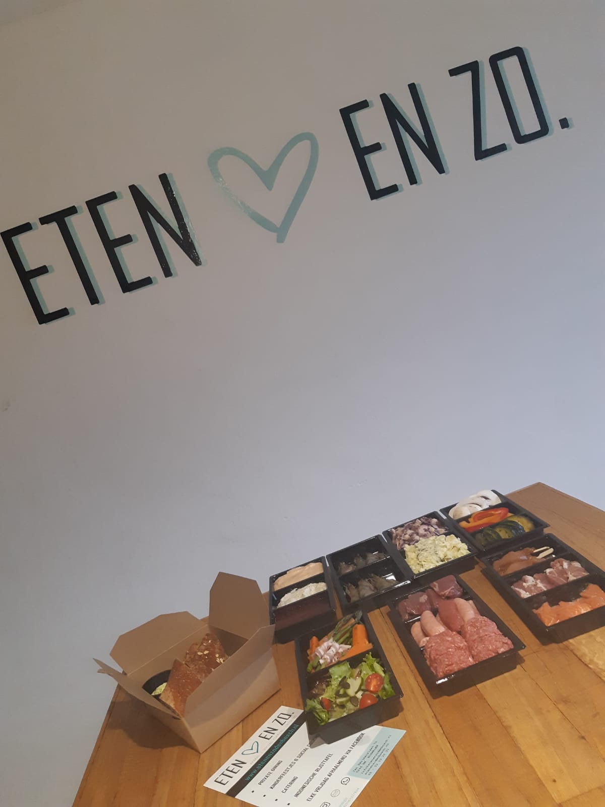 Meerdere voedselcontainers en een afhaaldoos op een houten tafel onder een muur met de tekst 