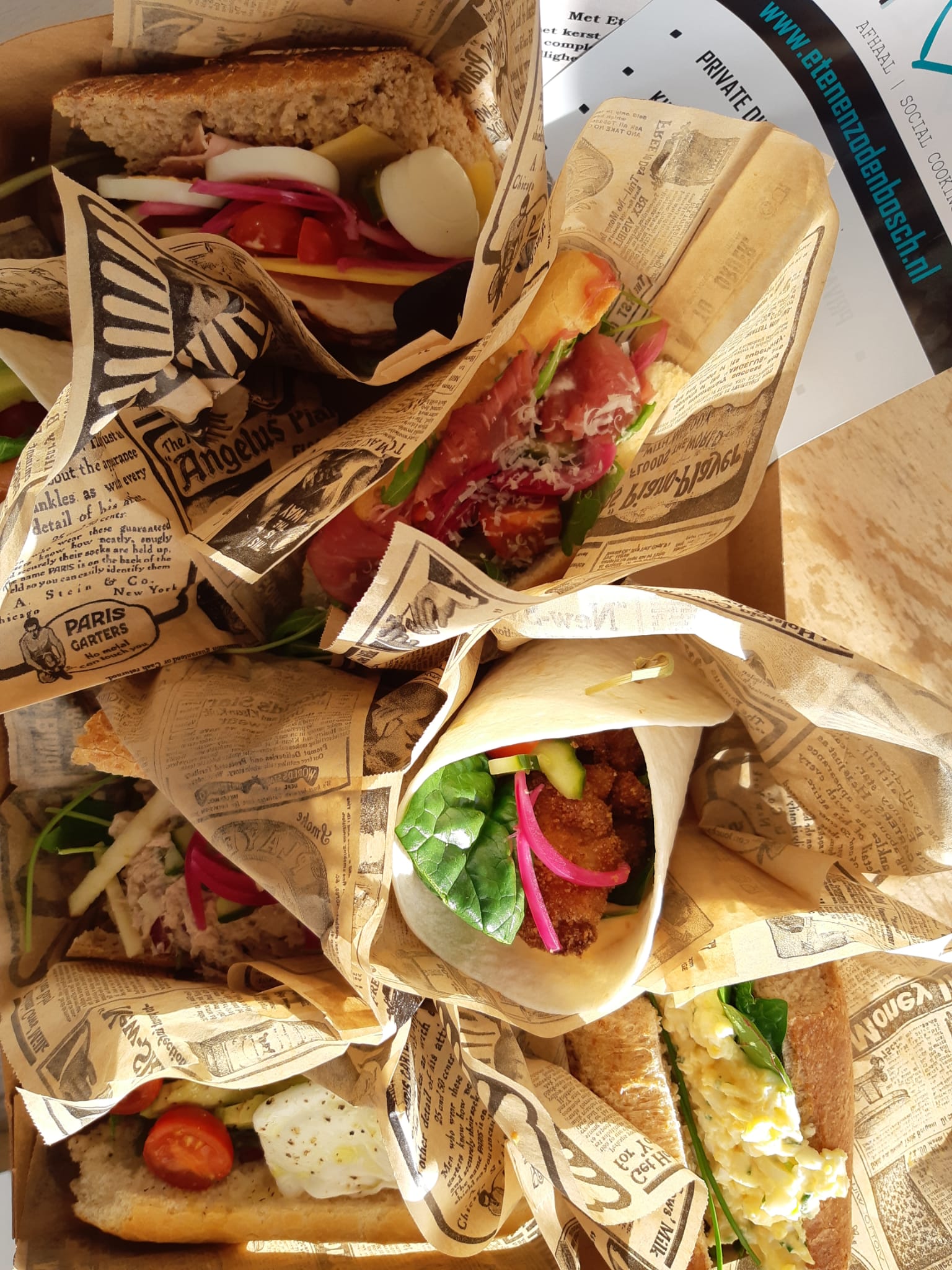 Vijf luxe sandwiches en wraps in bedrukte papieren verpakkingen, gerangschikt in een kartonnen doos.