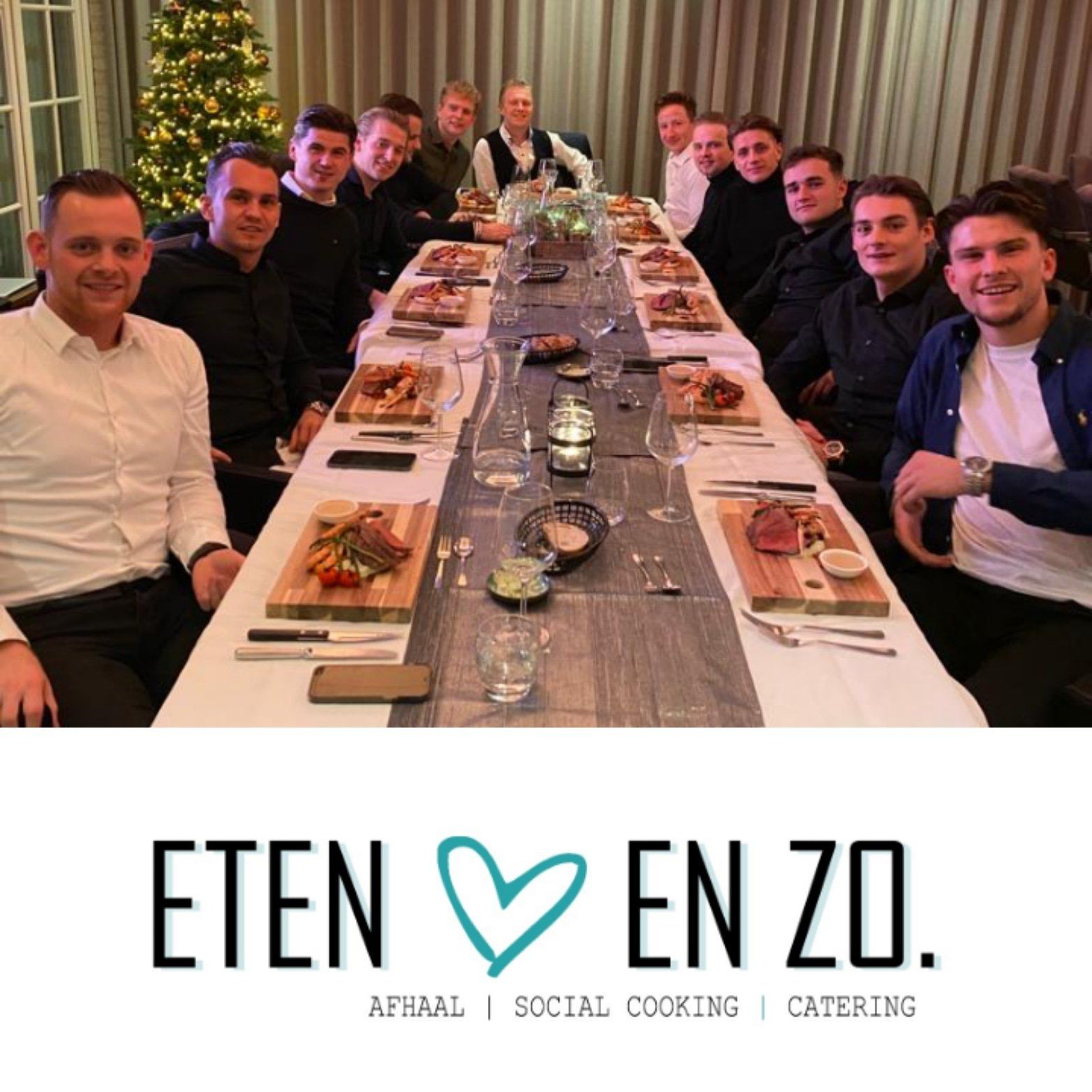 Een grote groep mensen zit aan een lange eettafel voor een feestelijke maaltijd, met daaronder de tekst 