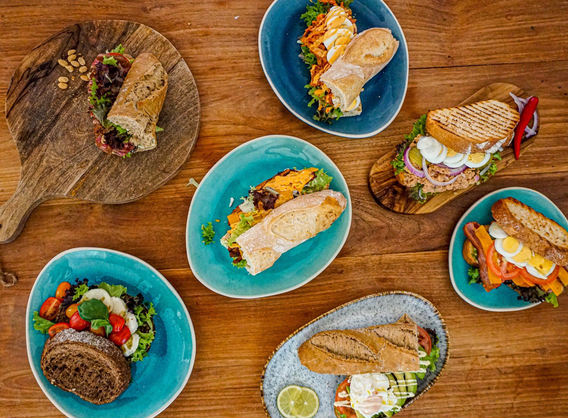 Bovenaanzicht van zes verschillende luxe sandwiches en salades, geserveerd op blauwe borden en houten planken op een houten tafel.