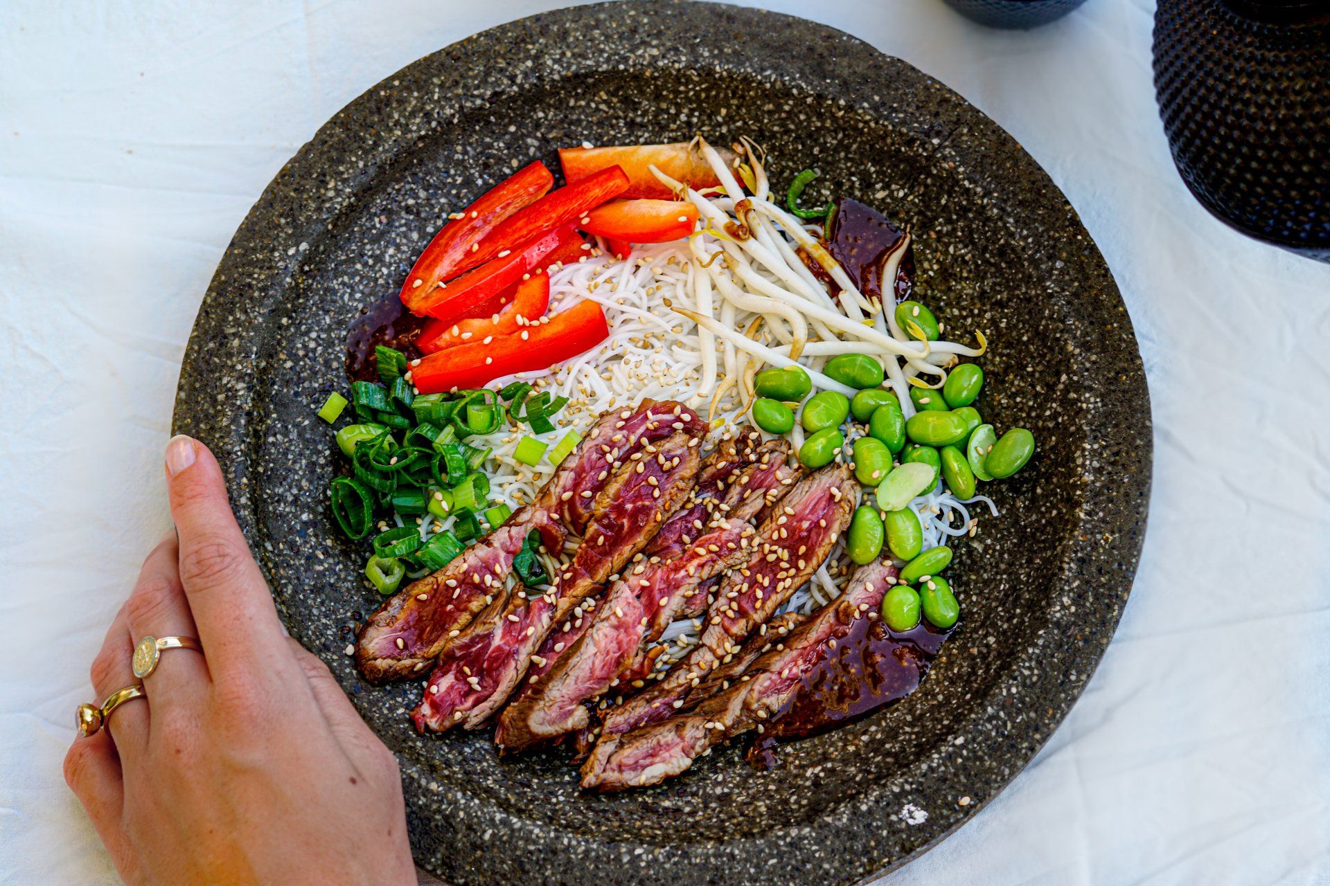 Een hand houdt een rustieke stenen kom vast, gevuld met aangebraden plakjes rundvlees, rijst, rode paprika, taugé, edamame en lente-uitjes.