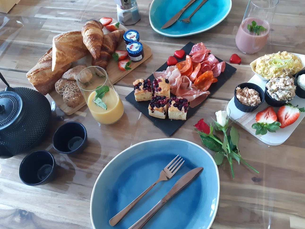 Een assortiment ontbijtproducten, waaronder gebak, vleeswaren, fruit en yoghurt, staat uitgestald op een houten tafel met blauw servies.