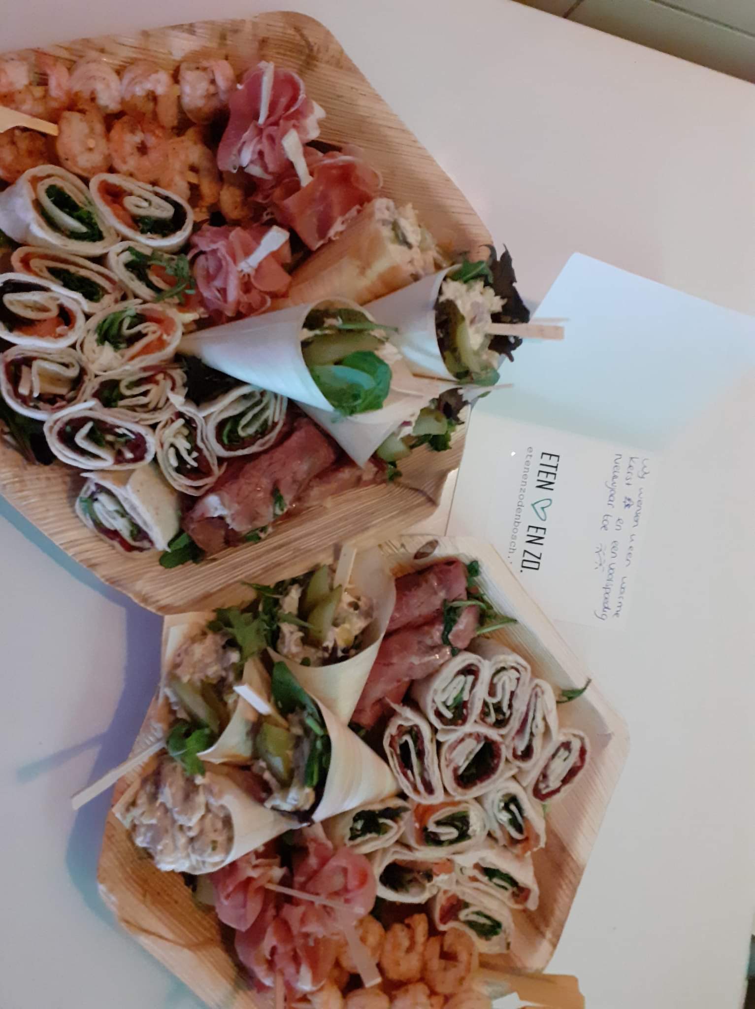 Twee houten bordjes met daarop rolvormige wraps, gevouwen crêpes, prosciutto en garnalen, en een klein briefje ernaast.