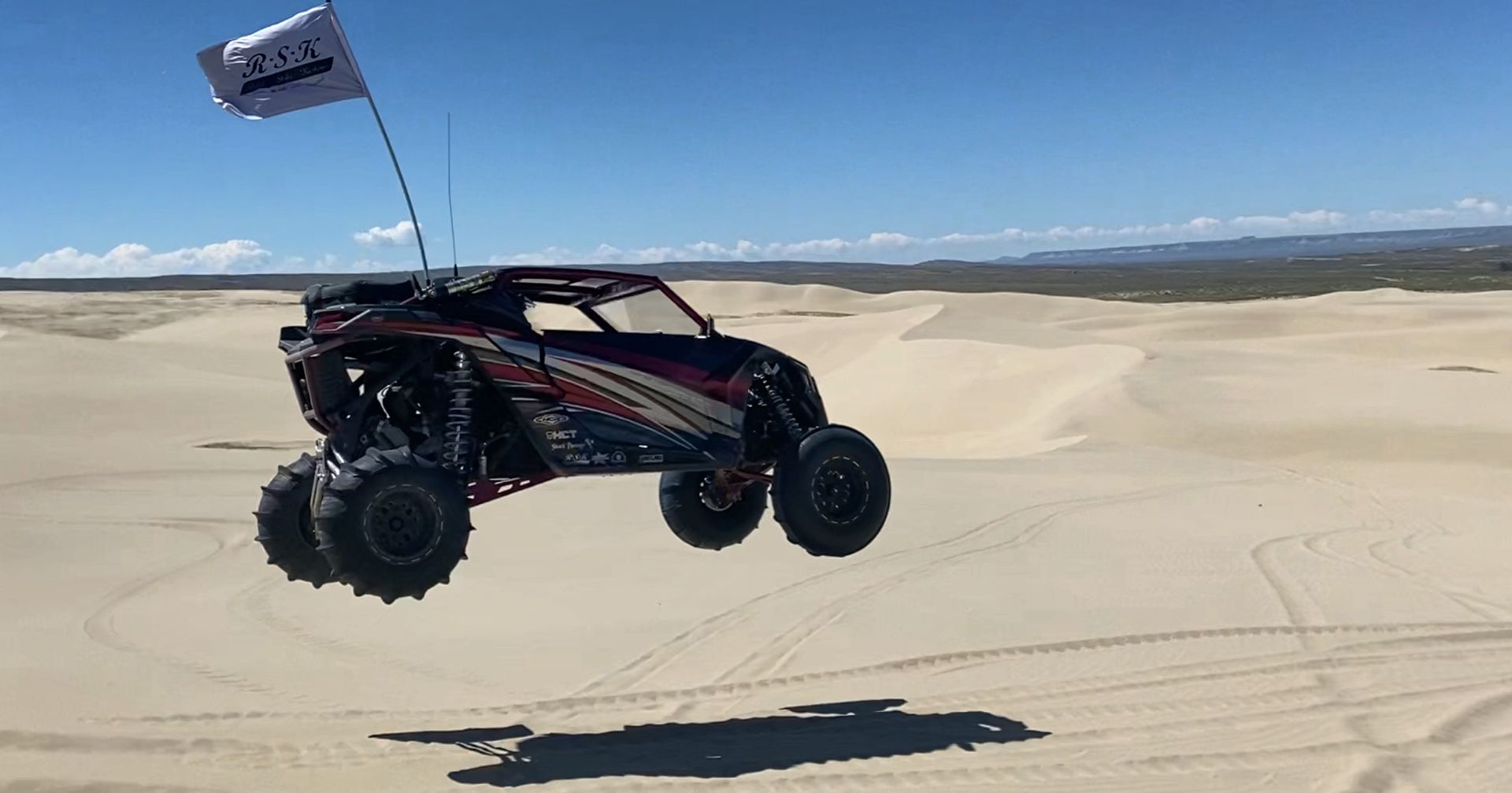 Side-by-side off-road buggy airborne over sand dunes with a flag waving behind it