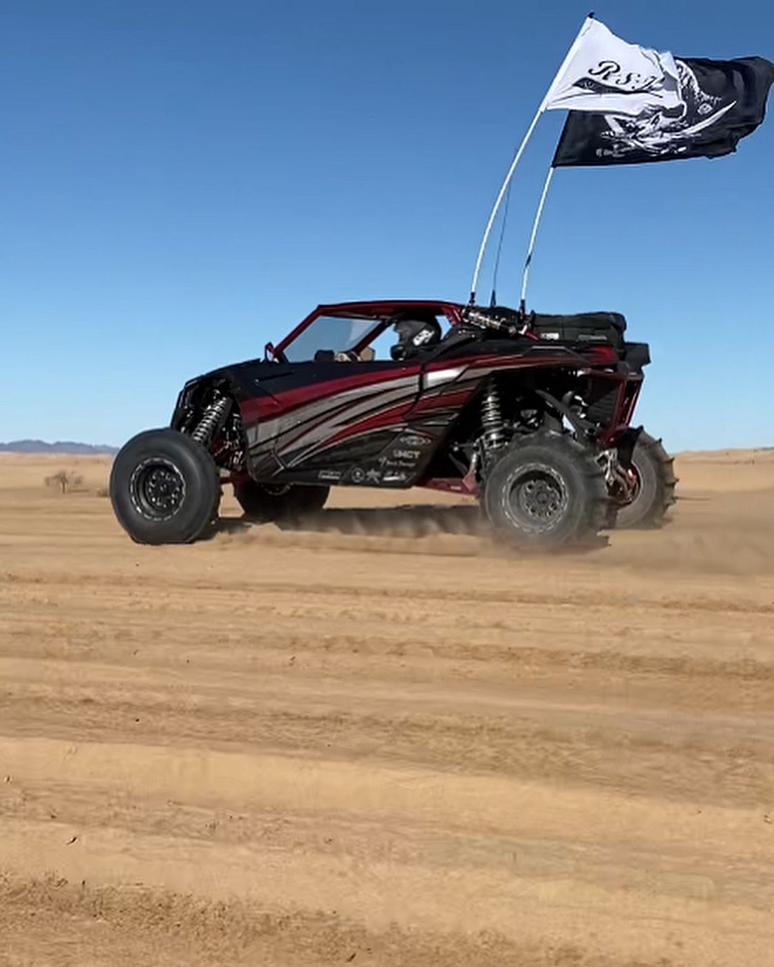Black off-road buggy driving fast across desert sand, flying a black-and-white flag under a blue sky