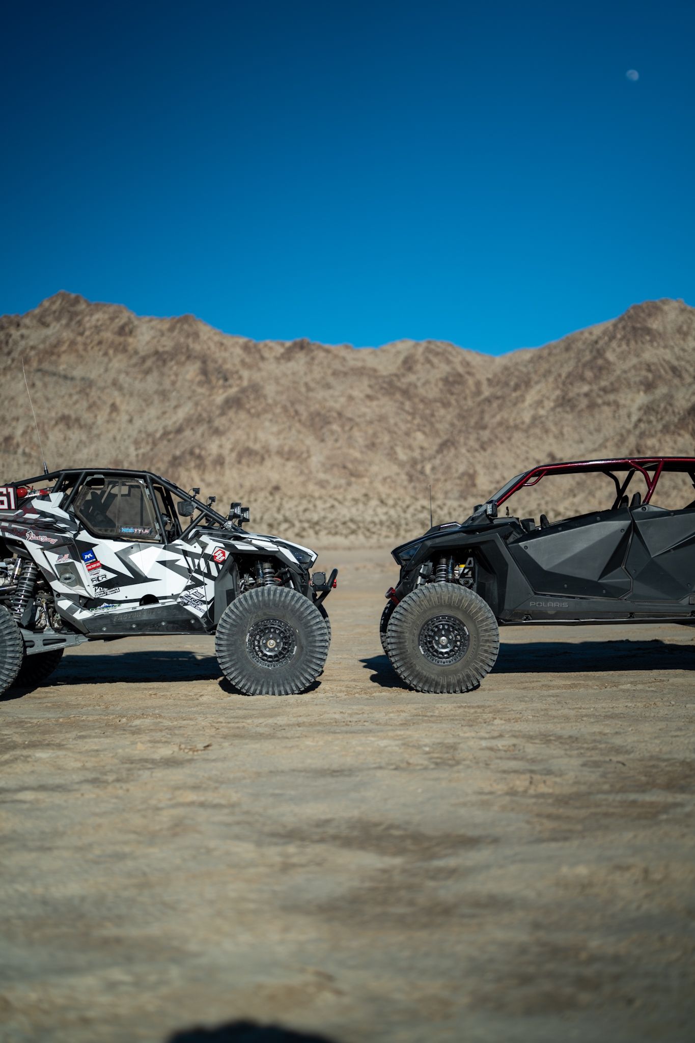 Two off-road UTVs parked on a dirt lot with tan hills and a clear blue sky in the background