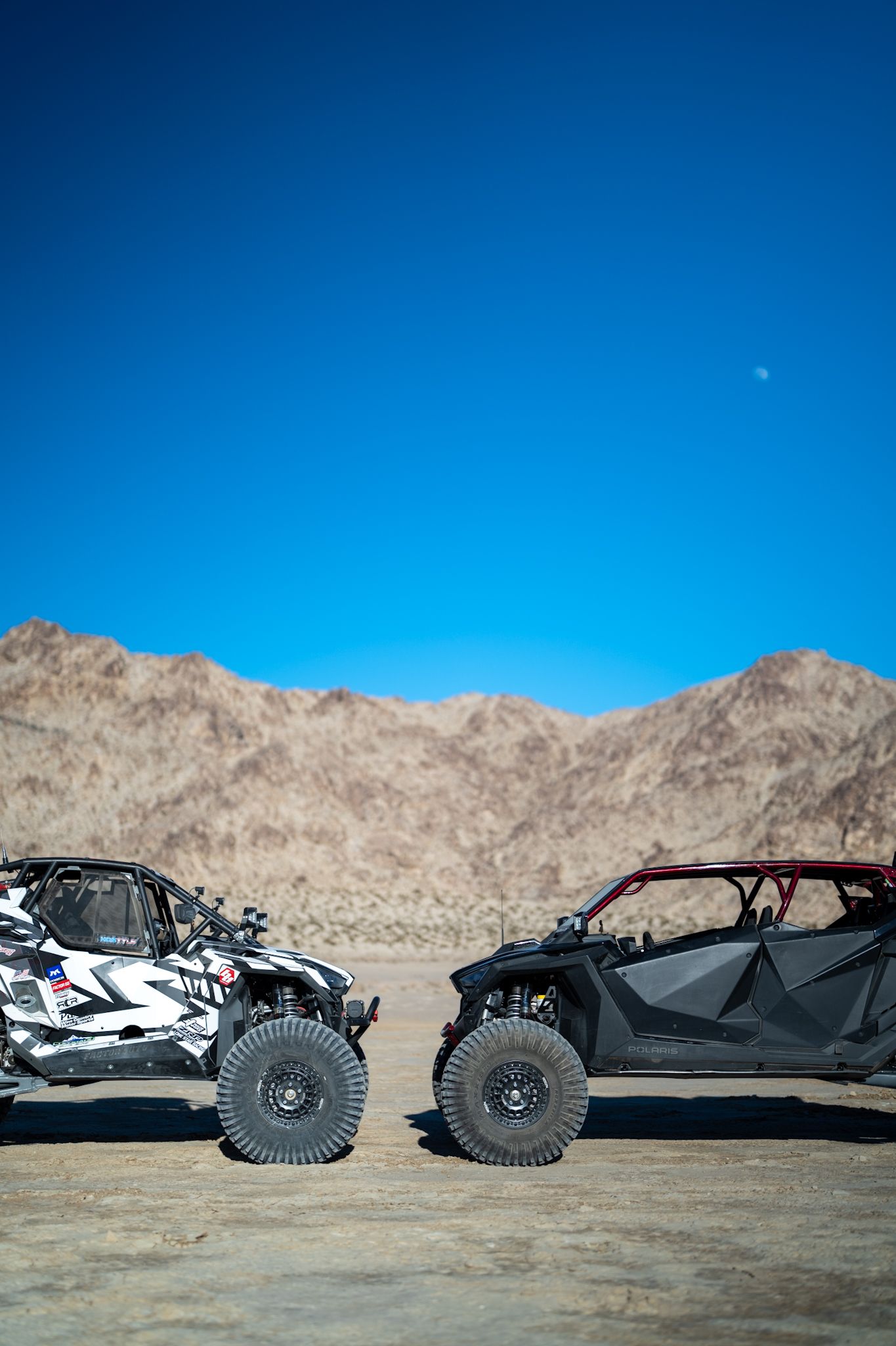 Two off-road dune buggies parked on a desert plain with mountains under a clear blue sky