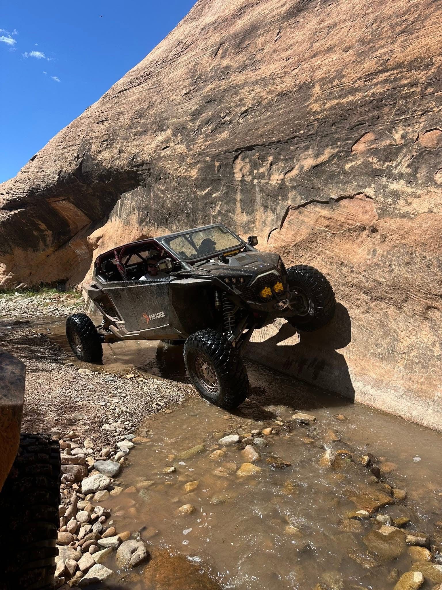 Off-road buggy crawling through a rocky creek beside a sandstone cliff under a blue sky