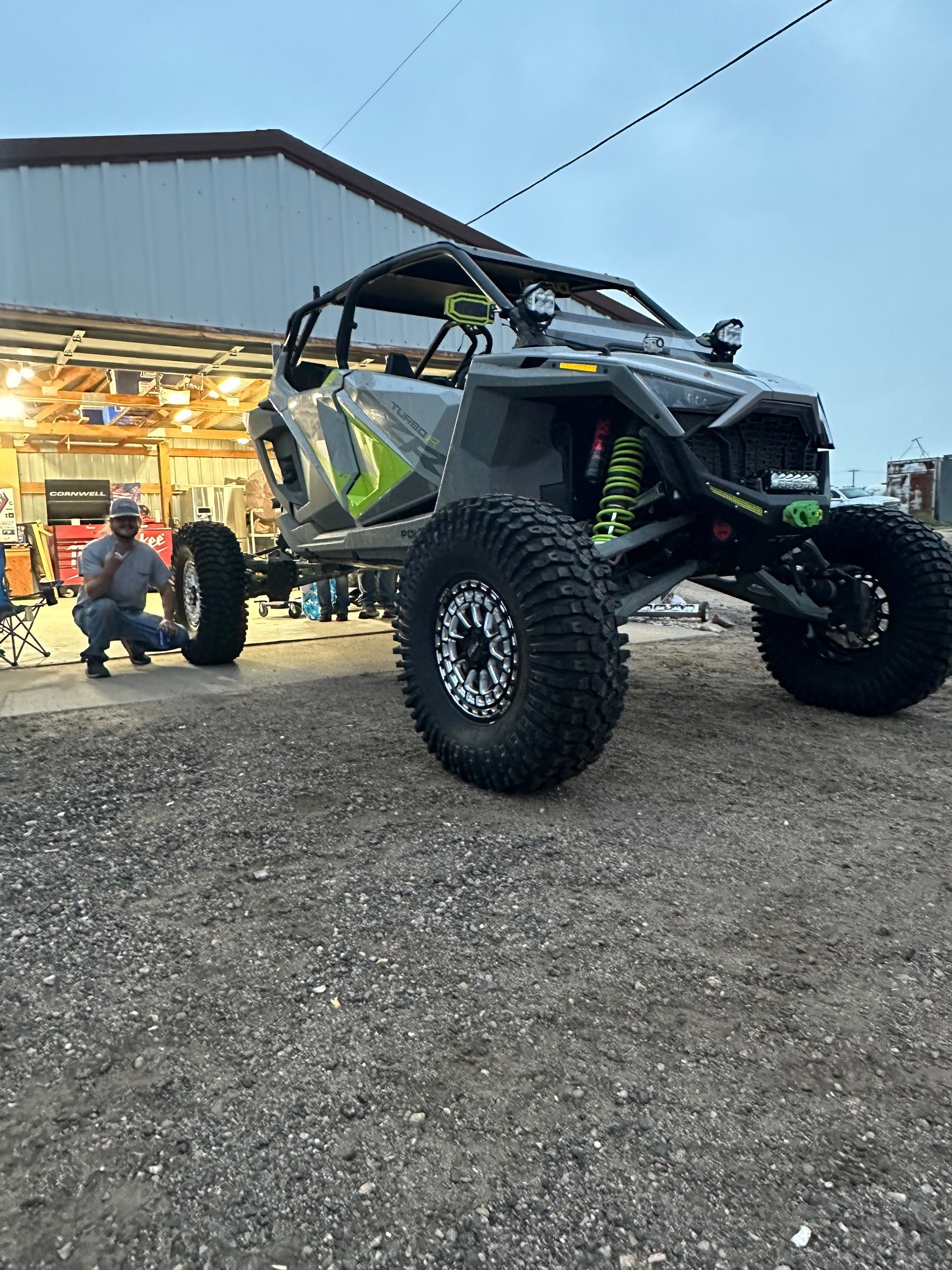 Off-road UTV parked outside a garage at dusk, with oversized tires and a person standing nearby