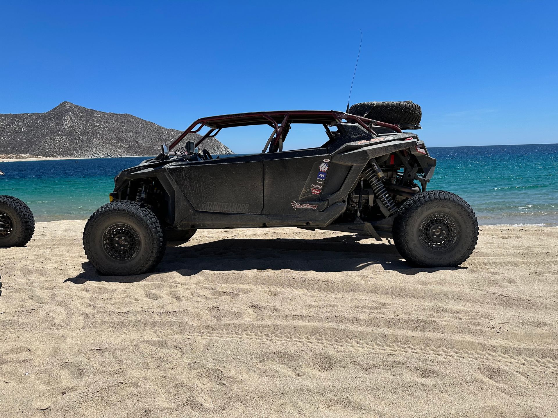 Black off-road dune buggy parked on a sandy beach with blue ocean and distant hills behind it