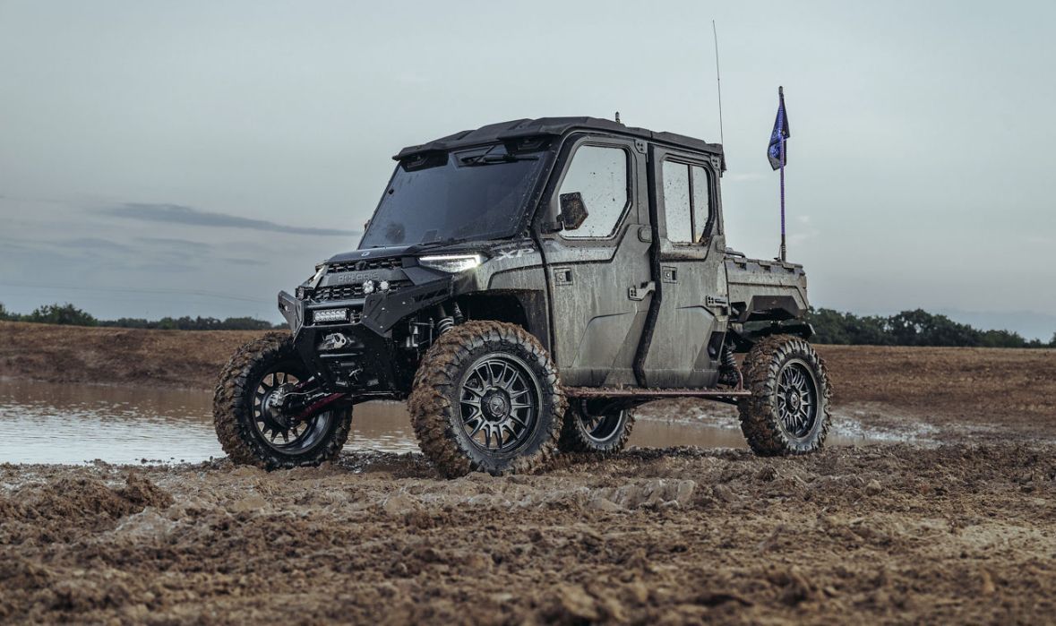 Muddy off-road vehicle parked on a wet dirt track under a gray sky, with a small flag mounted on the back