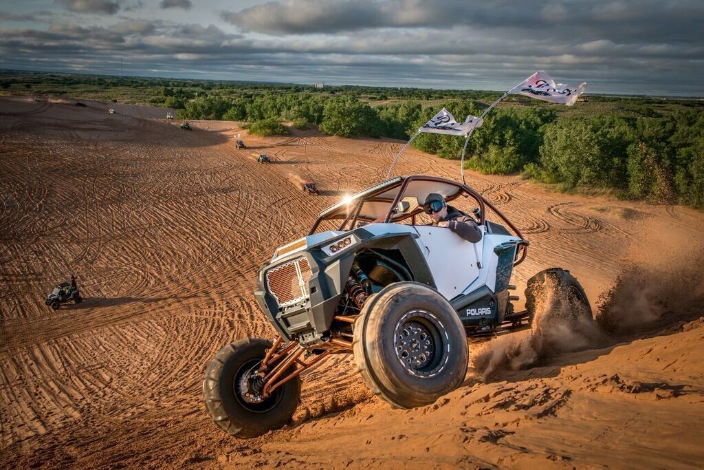 Off-road buggy racing across sandy dunes, kicking up dust under a cloudy sky