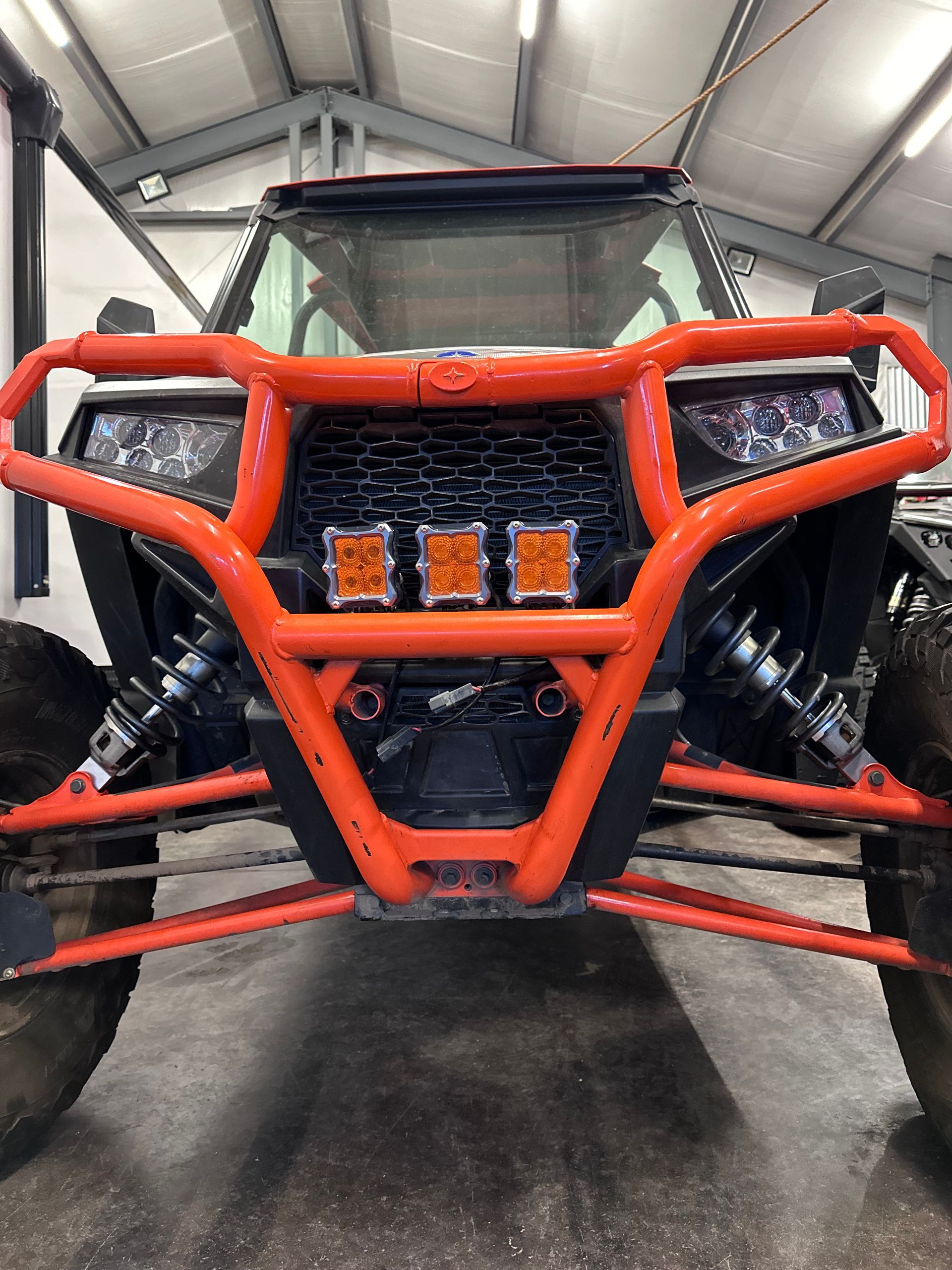 Front view of an orange off-road vehicle with a tubular bumper and four lights in a garage.