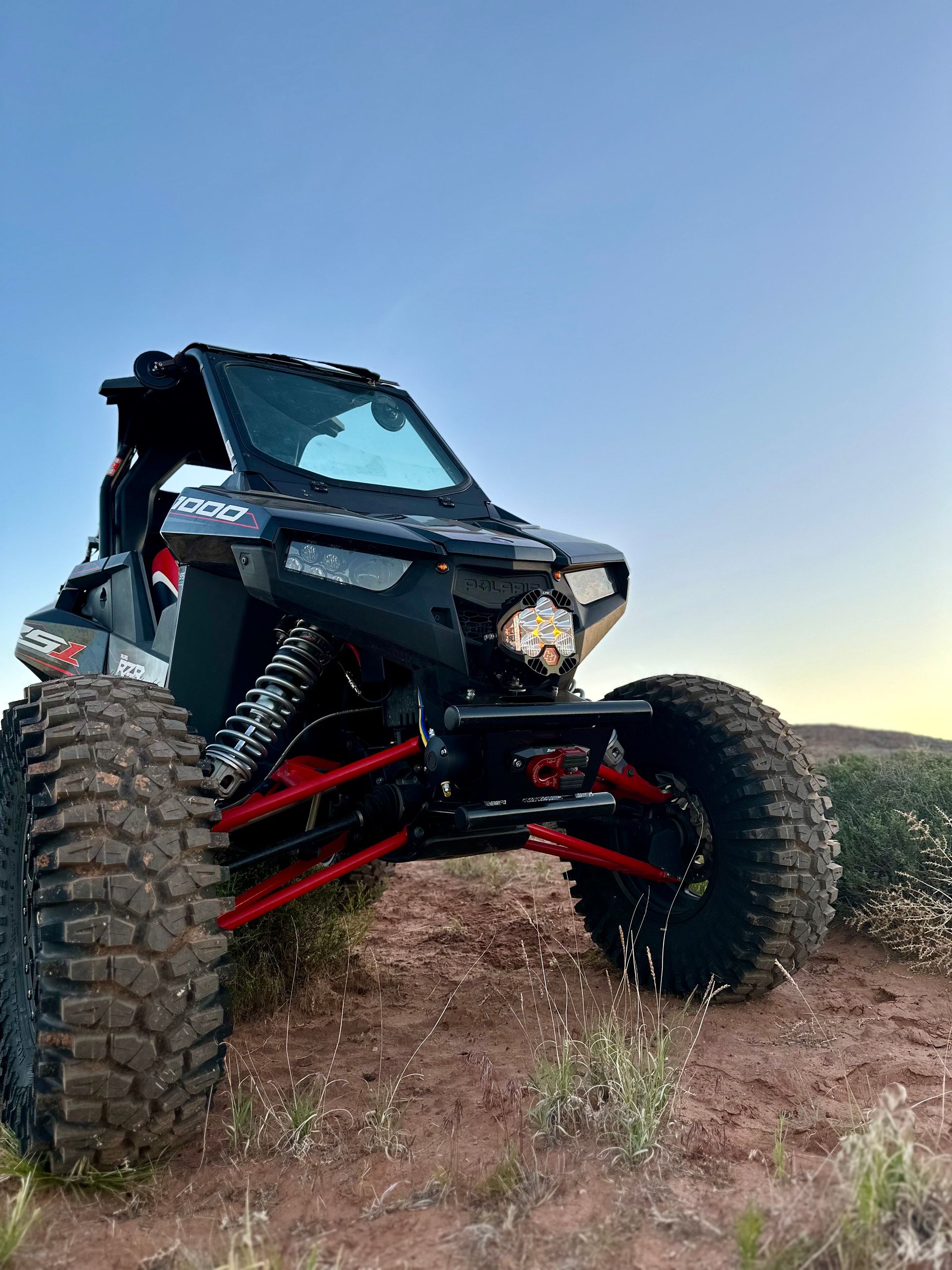 Black off-road side-by-side on a dirt mound at sunset with large knobby tires and red suspension