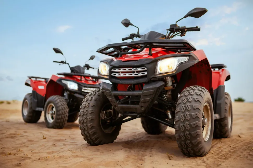 Two red ATVs on a sandy trail, front four-wheeler in focus against a blue sky.
