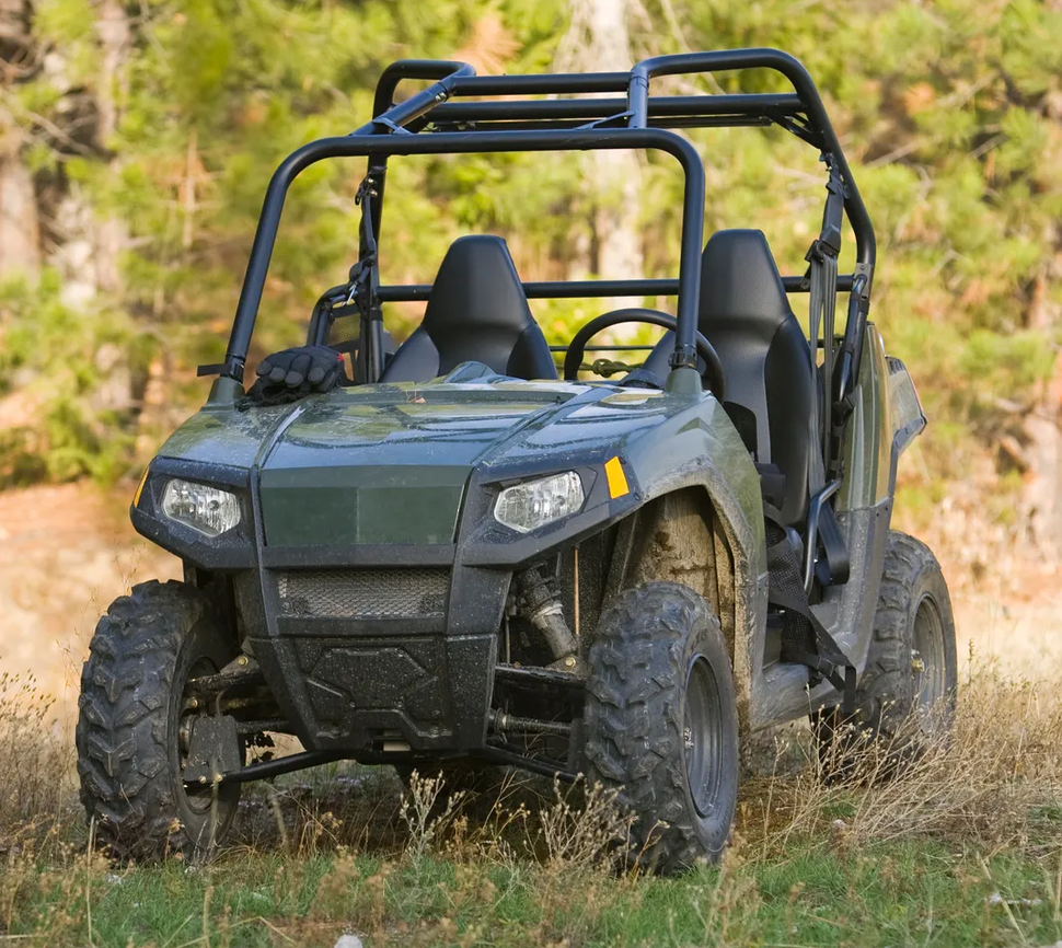 Green off-road utility vehicle driving through grassy woodland, front view with roll cage.
