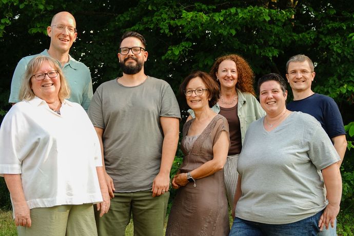 Sechs Personen posieren gemeinsam vor einer schlichten Wand in einem Innenraum und lächeln in Freizeitkleidung.