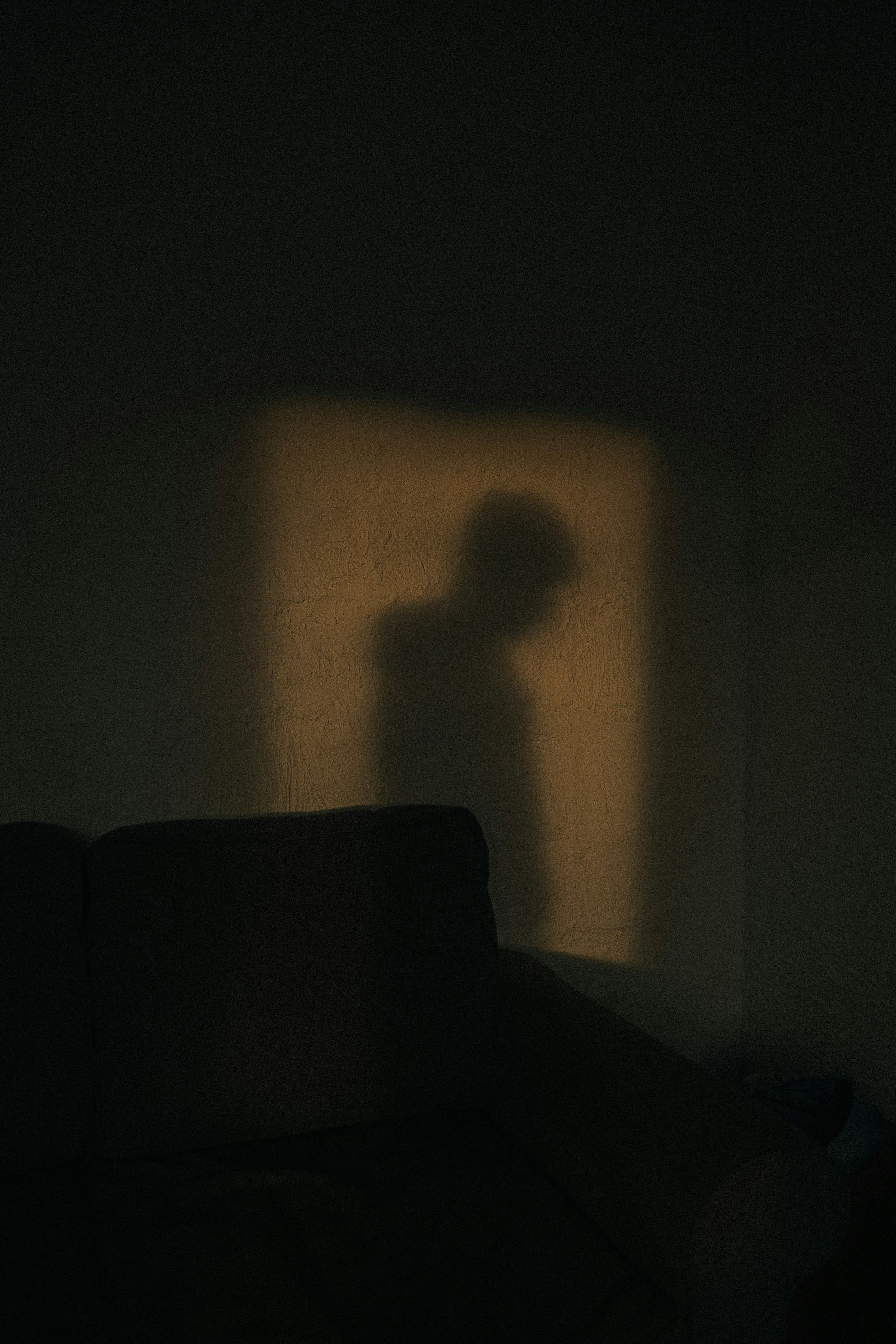 Ein schwach beleuchteter Raum, in dem der Schatten einer Person auf eine erleuchtete Wand hinter einem dunklen Sofa fällt.