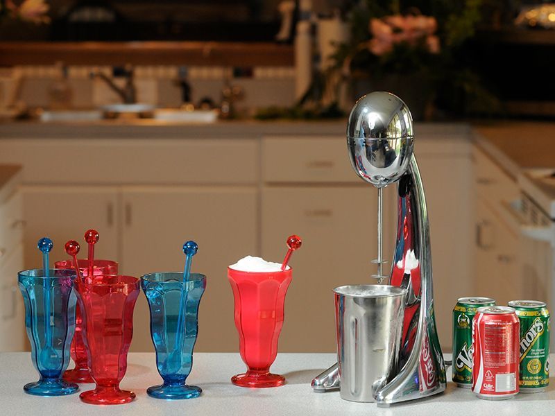 Milkshake maker with filled glasses, cans of soda, and colorful plastic cups on a countertop.