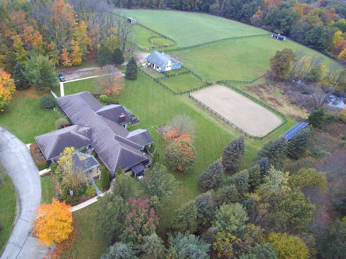 Aerial view of a large house with a long driveway and an adjacent equestrian area in a wooded area during autumn.