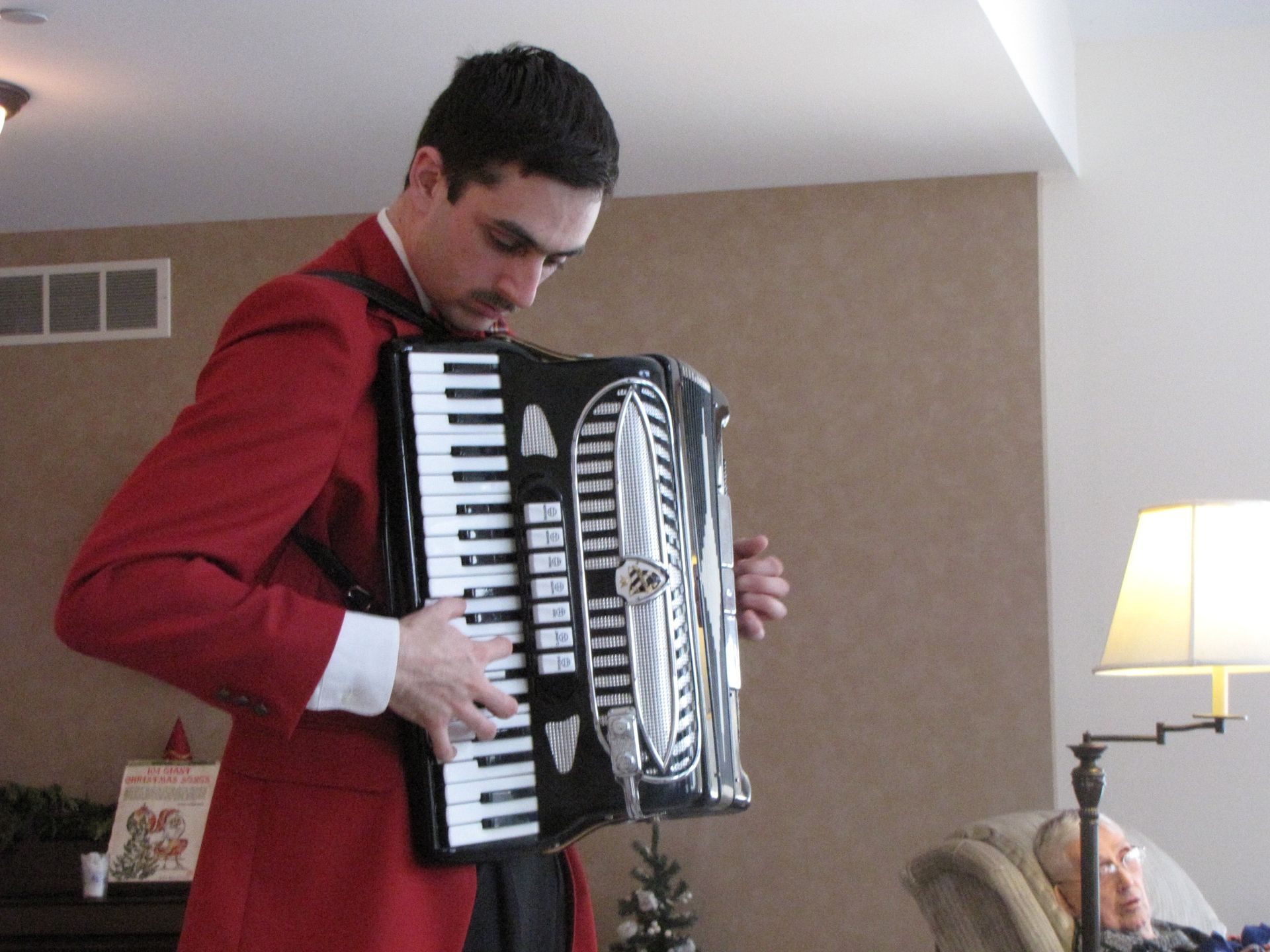 Man in red jacket plays accordion indoors near Christmas decor.