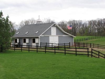 Gray stable horse barn with white trim and United States flag flying in front surrounded by dark brown wood pasture fencing and lush green grass.