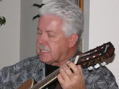 Man with gray hair and mustache playing a mandolin, eyes closed, wearing a patterned shirt.