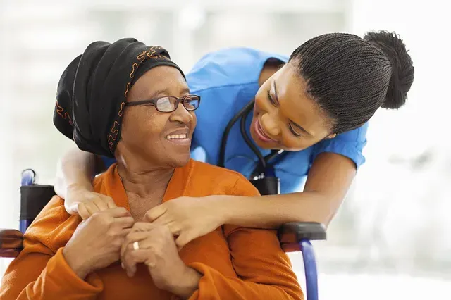 Nurse embracing patient in wheelchair, both smiling. The nurse wears blue scrubs, the patient wears orange.