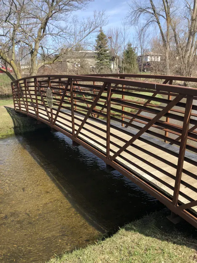 Brown metal pedestrian bridge over a shallow, clear stream in a park.