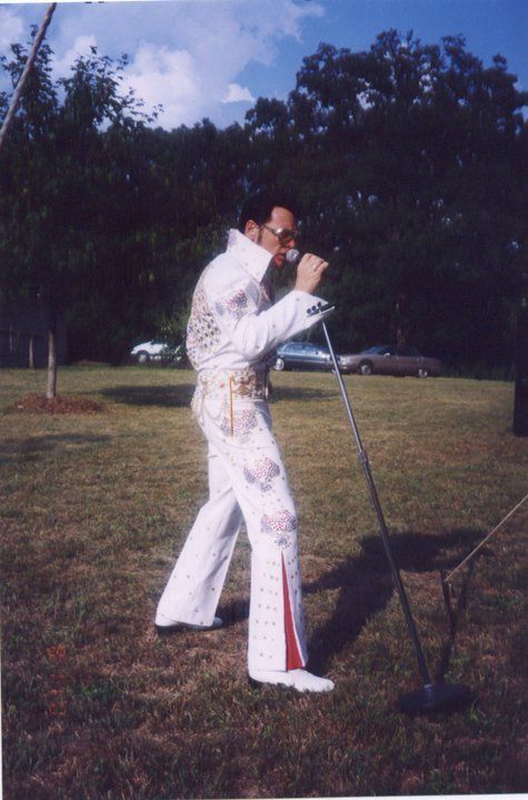 Man in Elvis costume singing into a microphone outdoors; trees in the background.