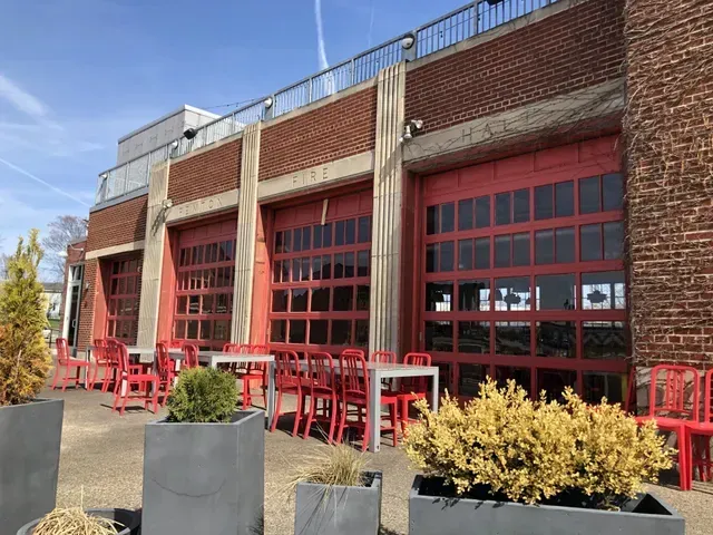 Exterior of a building with red garage doors. Outdoor tables and chairs are set up on a patio in front.