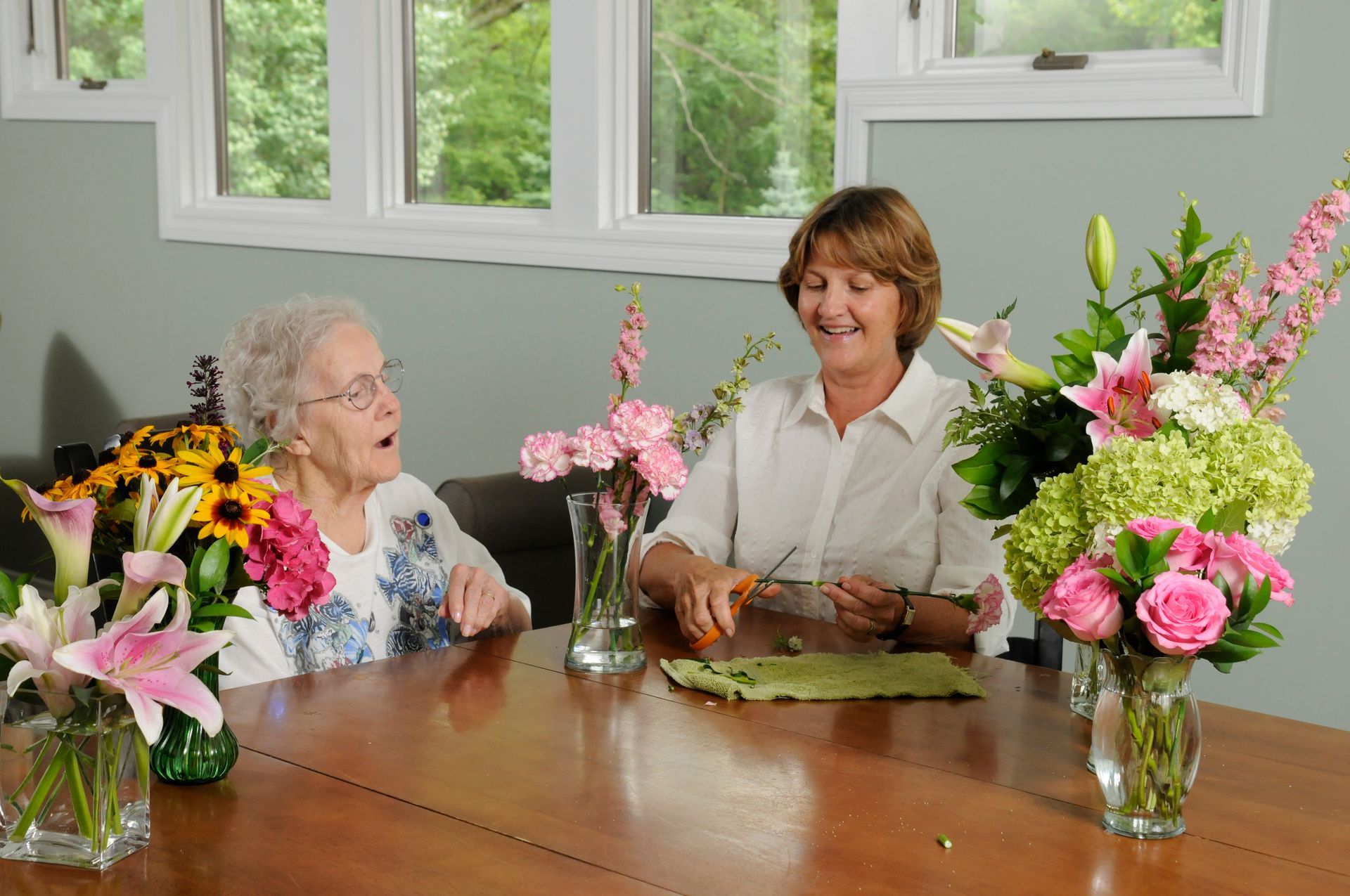 Woman and older adult arranging flowers at a table, smiling, with various colorful bouquets in vases.