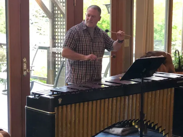 Man playing a xylophone with mallets; he's in a room with a large window.