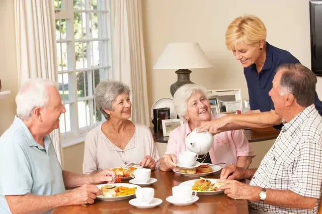 Caregiver pouring tea for a group of people seated at a dining table.