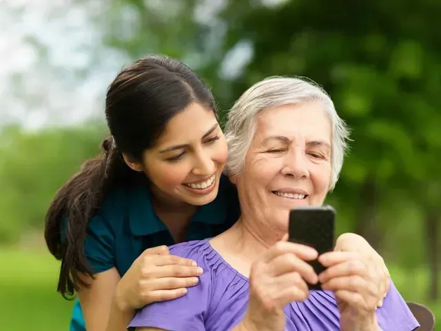 Woman showing elderly person a phone, both smiling outside.