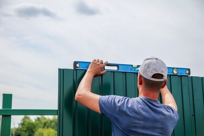 A hand uses a blue-bristled brush to apply a clear wood stain to a light-colored wooden fence board.