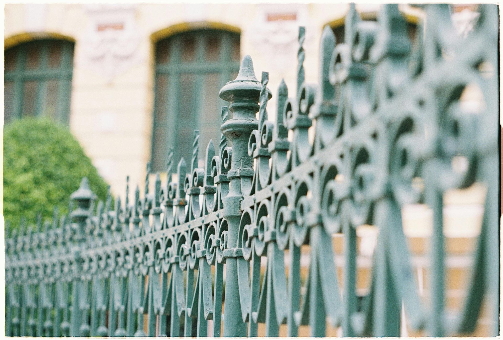 A shallow-depth-of-field view of an ornate, teal-green wrought iron fence in front of a blurry building.