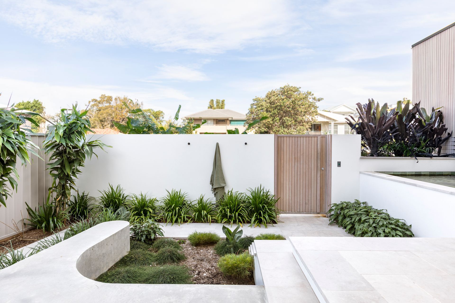 Modern, minimalist garden with white walls, wooden gate, and various green plants under a cloudy sky — Escarpment Building in Austinmer, NSW
