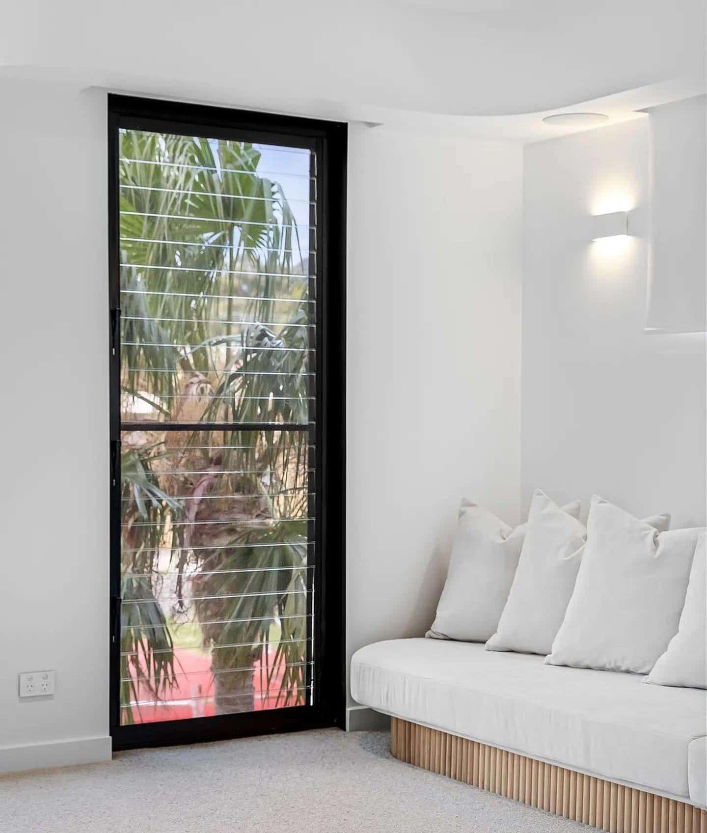 Black-framed glass door, white walls, and a white sofa with pillows. Palm tree visible through the glass — Escarpment Building in Austinmer, NSW
