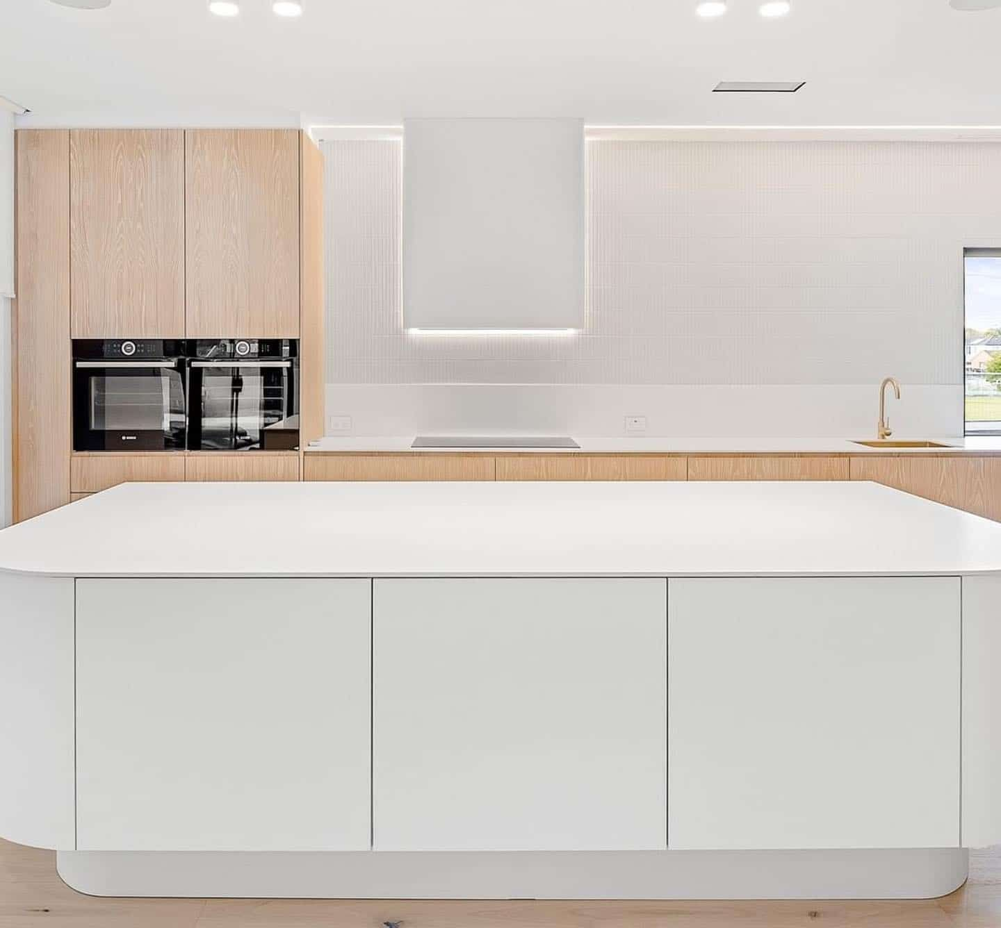 Modern white kitchen with a large island, wood cabinets, and built-in ovens — Escarpment Building in Austinmer, NSW