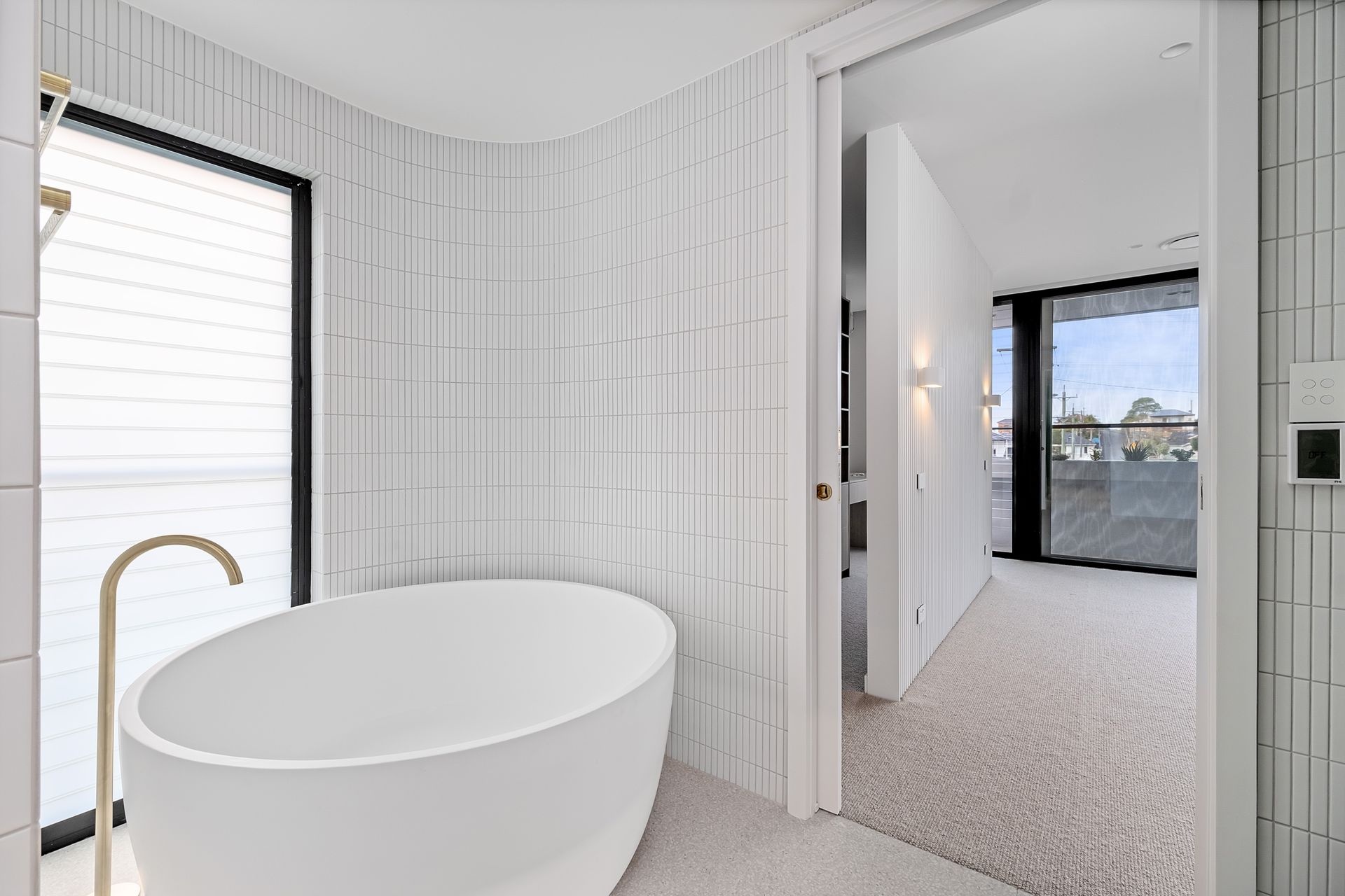 Modern white bathroom with a tub, gold faucet, and textured tile. Doorway to hallway — Escarpment Building in Austinmer, NSW