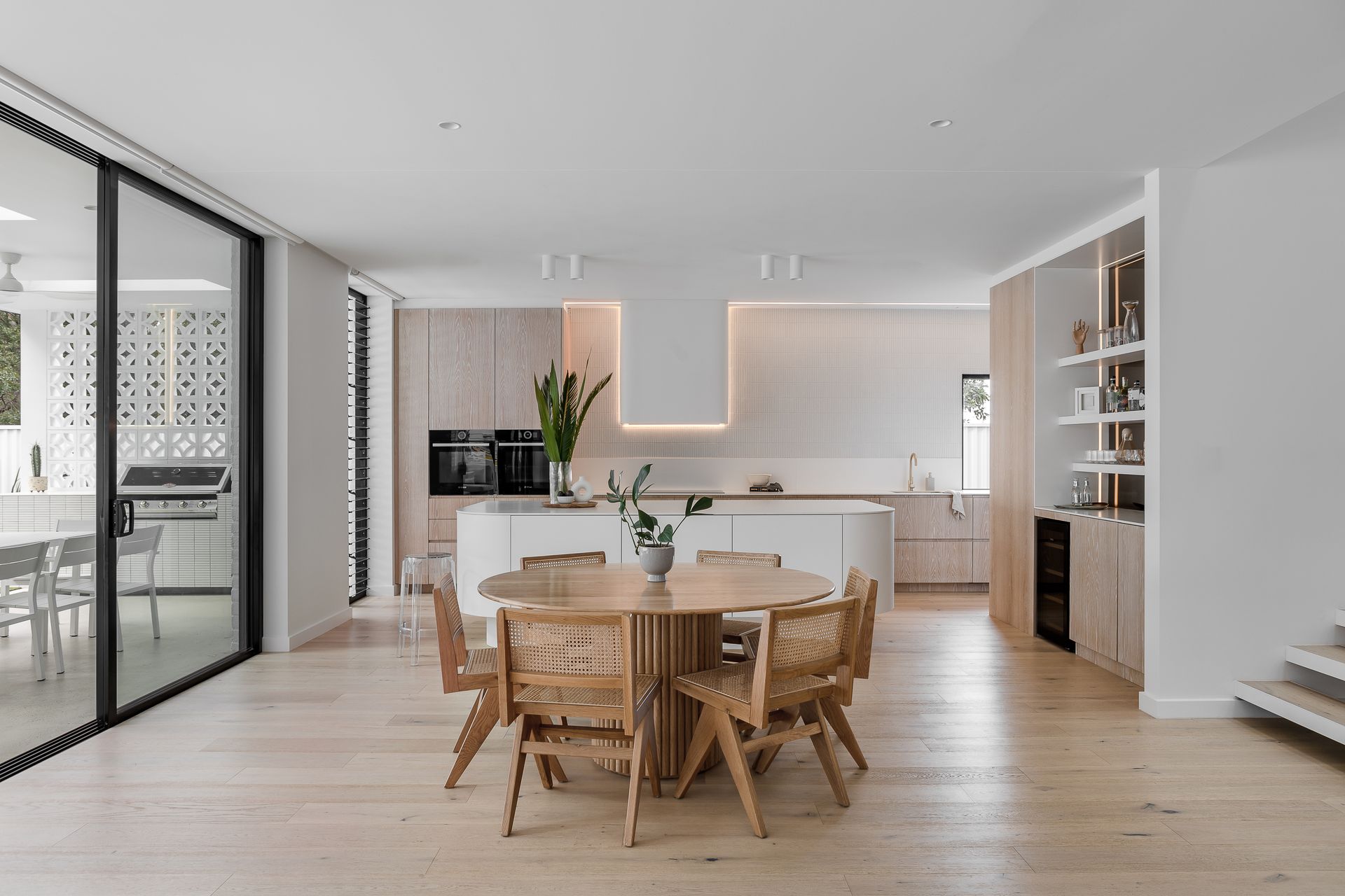 Open-plan kitchen and dining area with light wood floors, cabinets, and a round wooden table with woven chairs — Escarpment Building in Austinmer, NSW