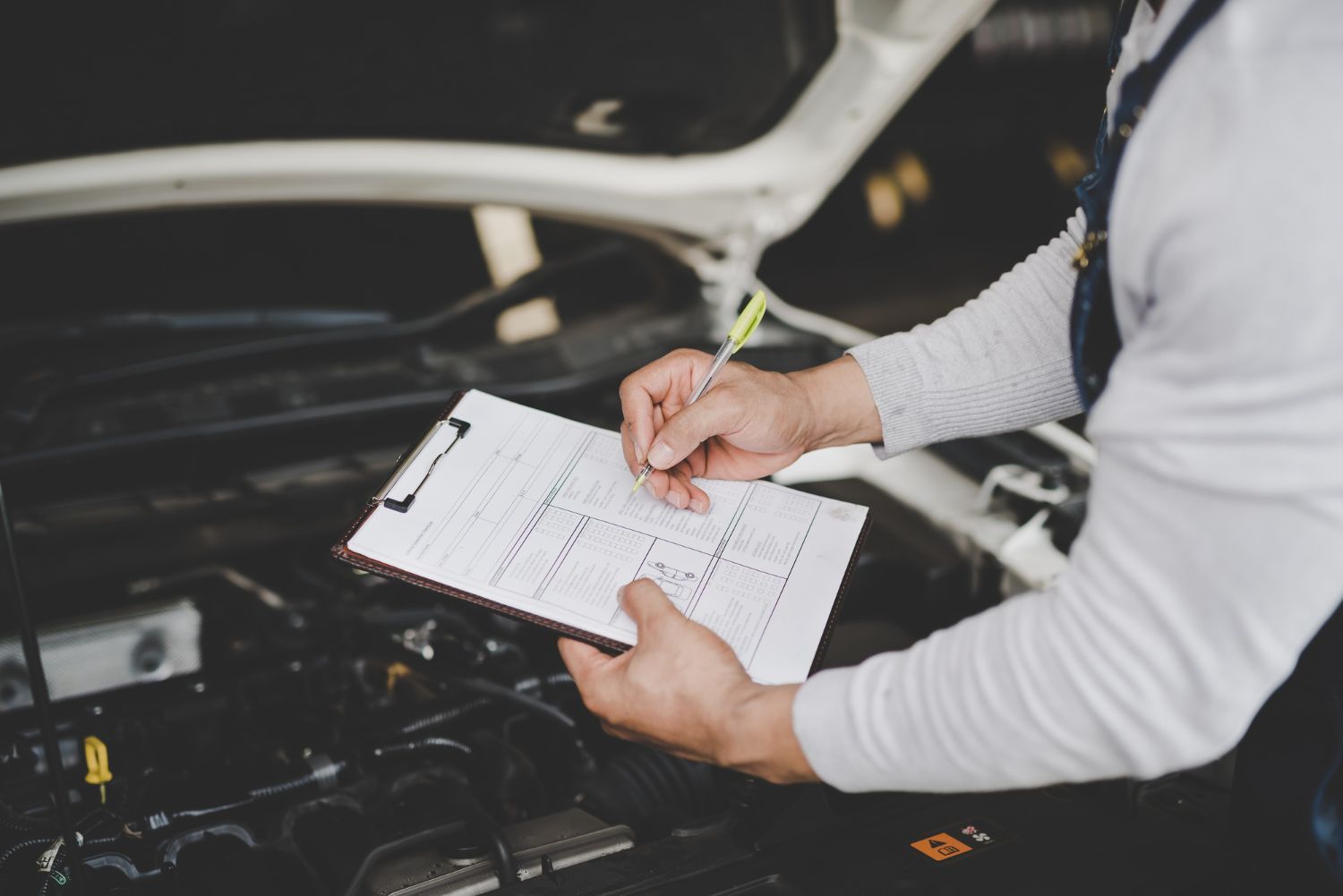 Mechanic writing on a checklist under a car hood in a garage