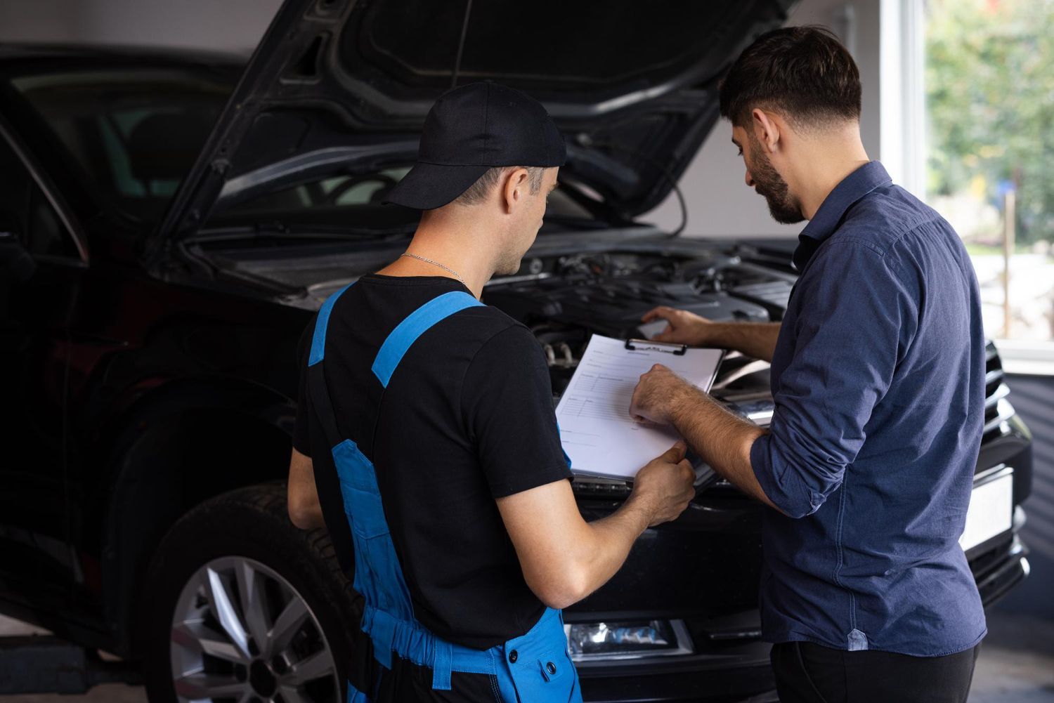 Two mechanics inspecting a car engine in a garage, with one holding paperwork.