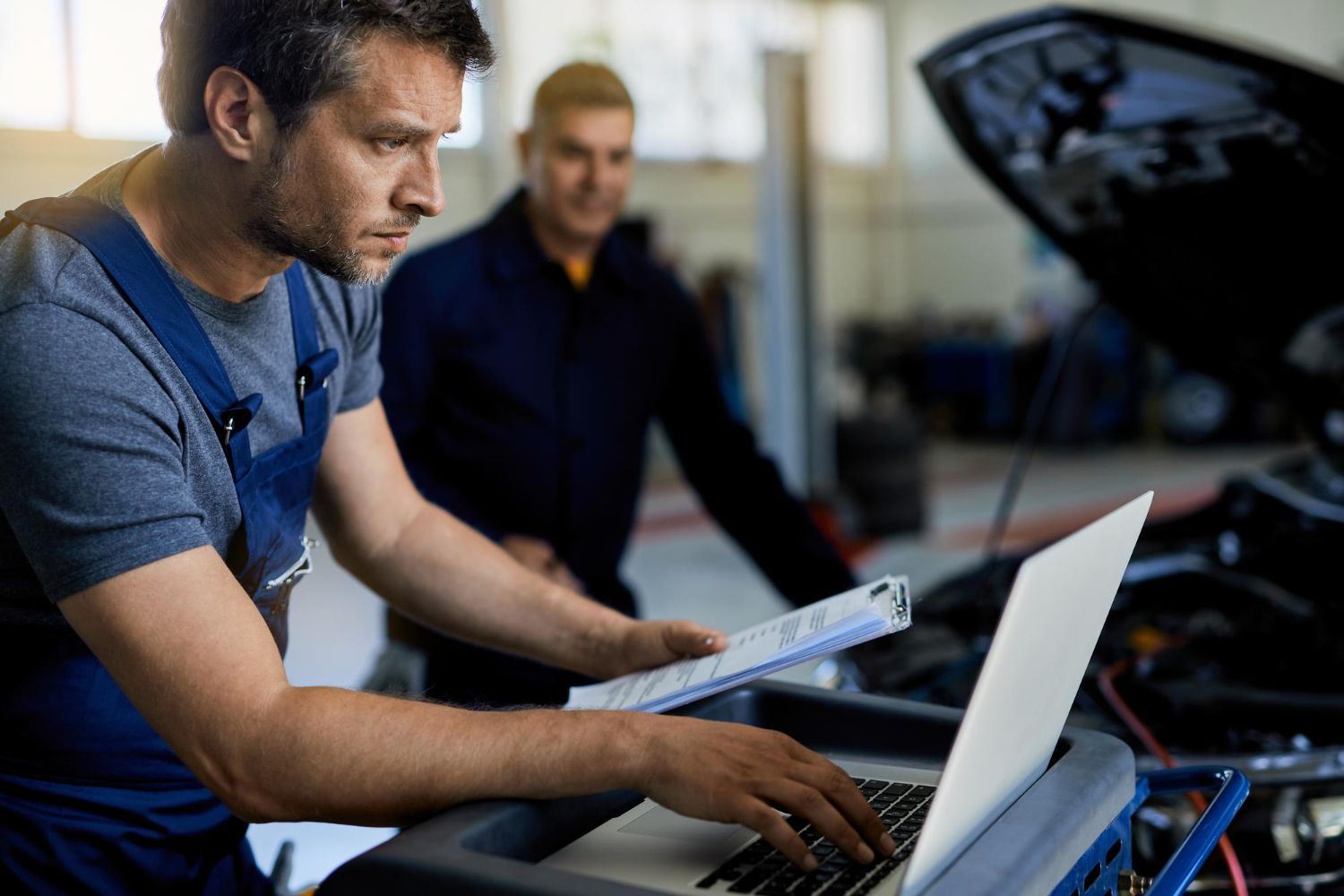 Technician working on a laptop beside an open car hood in a workshop