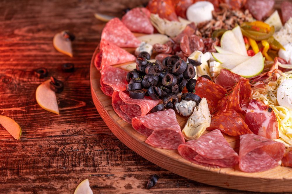 Tabla de embutidos: Carnes cortadas en rodajas, aceitunas, queso y rodajas de pera sobre una superficie de madera.