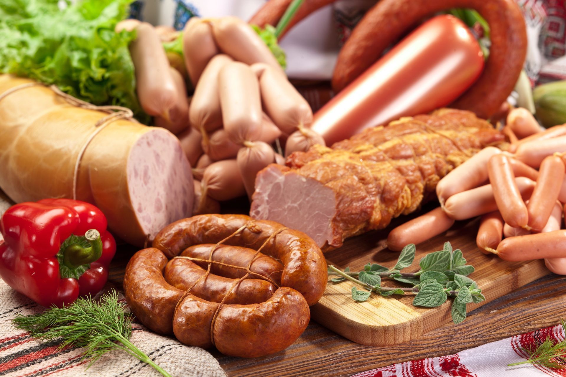 Surtido de embutidos y productos cárnicos, incluyendo espirales, salchichas y jamón en lonchas, con pimiento rojo y hierbas.