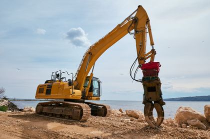 Yellow excavator digging on a rocky shoreline beside calm water under a cloudy sky