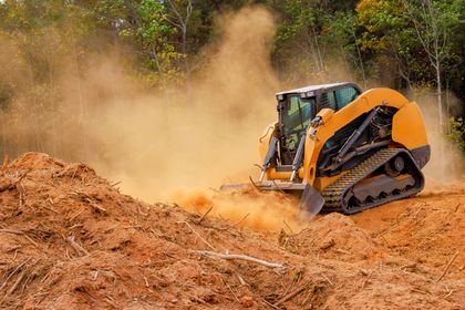 Orange skid-steer loader digging in a dirt field, kicking up dust near trees